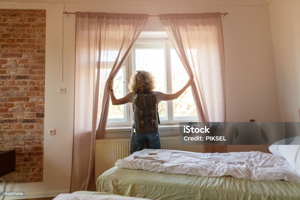 Rear view of tourist in the hotel room pulling the curtains to see the view Dorm Room Stock Photo