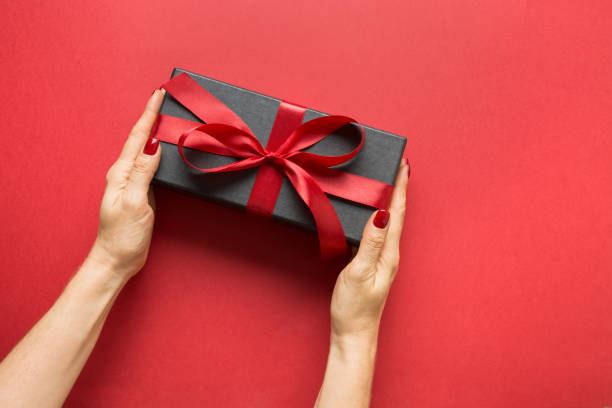 istock Woman's hands holding black gift box wrapped with red ribbon on red surface. Valentine day card. 1203776250