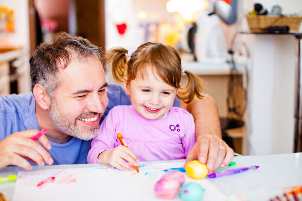 istock Portrait of Smiling Dad and Daughter Sitting at Table Painting Easter Eggs with Markers in Kitchen - stock photo 1203777708