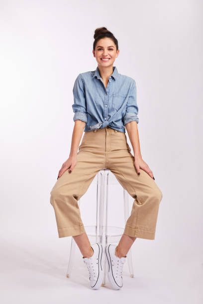 Smiling young brunette woman sitting on a stool on gray Studio portrait of a smiling young brunette woman wearing casual clothing sitting on a stool against a gray background sitting stock pictures, royalty-free photos & images