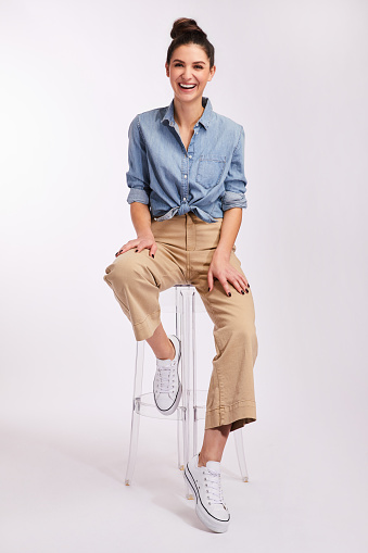 Studio portrait of a casually dressed young brunette woman laughing while sitting on a stool against a gray background