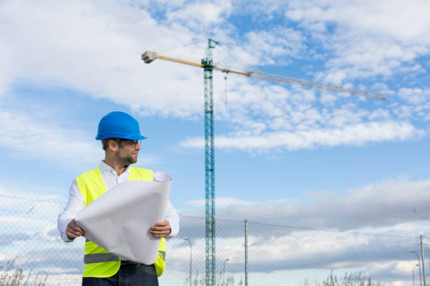 istock Architect or Engineer man holding blueprints on the Construction Site. Wearing protection clothes. Crane on background over blue sky. Job concept. Back light 1203798447