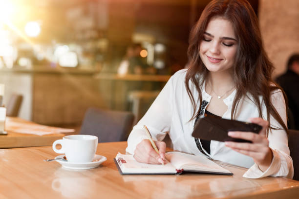 istock young beautiful woman taking notes to her planner using time schedule from her smartphone drinking hot coffee in the cafe multitasking modern businesswoman 1203803284