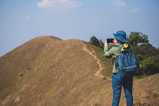 The back view of asian woman traveler backpack And wearing a jean hat Taking pictures of the mountain with a mobile phone