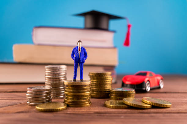 miniatiure people: a businessman standing on stacks of cons in the front of books, a graduation hat and a cute red car on a light blue backround. saving money, finance, business, education concept. - university graduation car student imagens e fotografias de stock