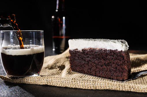 Portion of chocolate cake and black beer on black background, pouring beer into glass