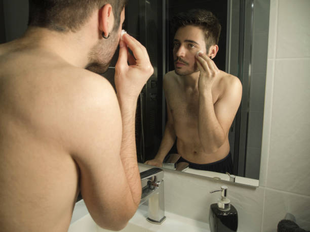 portrait of handsome caucasian man reflection in from of the mirror, cleaning his face with cotton make-up removers - shaved head imagens e fotografias de stock