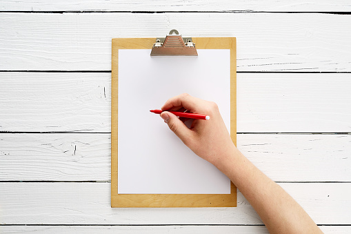 Hand with red pencil is writing on a piece of paper on a clipboard. Clipboard is empty and on a white background.