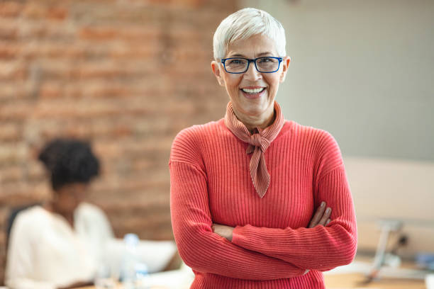 Mature female in office with team meeting in background. Close-up portrait of mature business woman standing at office. Pretty older business woman, successful confidence with arms crossed in financial building. Mature female in office with team meeting in background. female role model stock pictures, royalty-free photos & images