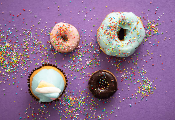 Delicious doughnuts and cupcake with sprinkles on purple color background, top view
