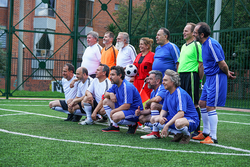 two soccer teams made up of older Latino men divided into two groups the blue uniform team and the white uniform team and a female referee are organized for a photo six players two of the white team two archers two of the blue team and the referee of skin in the back and six players three of the white team and three of the blue team in the front kneeling all are dento the court and look at the camera