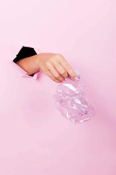 istock Female hand holds through a hole a empty crumpled plastic bottle on a pink background, no plastic and zero waste concept 1203834134