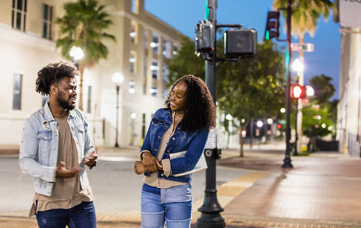 A young African-American couple hanging out in the city, talking  and smiling as they walking across the street. It is evening, with the street lights illuminated.