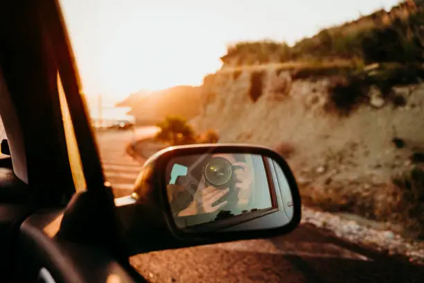 Photo of woman taking a self portrait in rear mirror in a car at sunset. travel concept
