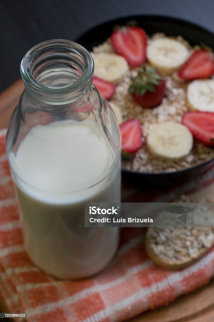 milk bottle with bowl of oatmeal and fruits background Close-up of a bottle with milk and in the background a bowl with oatmeal and slices of strawberries and bananas with a wooden spoon with oatmeal in the background Argentina Stock Photo