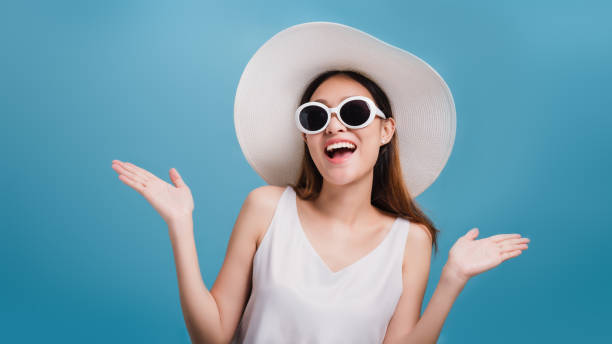 アジアの笑顔美しい女性ブロガーは広い帽子をかぶり、コピースペースで青い色の背景に隔離されたサングラスを着用しています。プロモーションと販売と旅行ビジネスのコンセプト。 - hat shop ストックフォトと画像