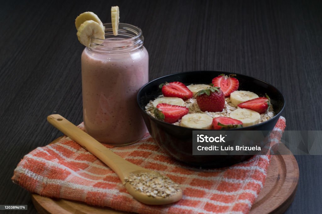 Banana strawberry and oatmeal smoothie with its ingredients Banana, strawberry and oatmeal smoothie in a jar with slices of bananas and strawberries in a bowl full of oatmeal and next to a wooden spoon with oatmeal, top of a rag with a round wooden down Argentina Stock Photo