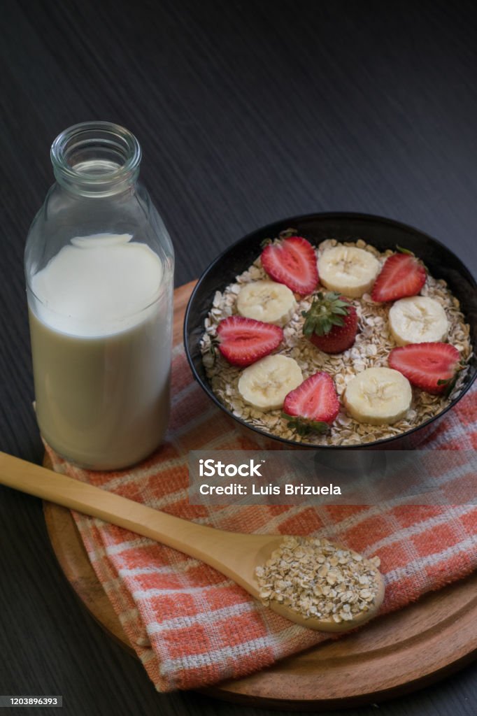 Bowl with oatmeal and fruits, milk bottle and wooden spoon with oatmeal Medium shot of a bowl with oatmeal and slices of banana and strawberry with a bottle with milk and a wooden spoon with oatmeal Argentina Stock Photo