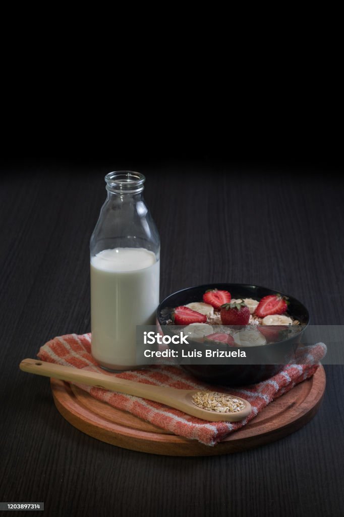 Bowl with strawberry and banana oatmeal with a bottle of milk general view of a bowl of oatmeal with a wooden spoon with two strawberries with a bottle of milk and bananas around and a jug of blender on top of a round wooden board Argentina Stock Photo