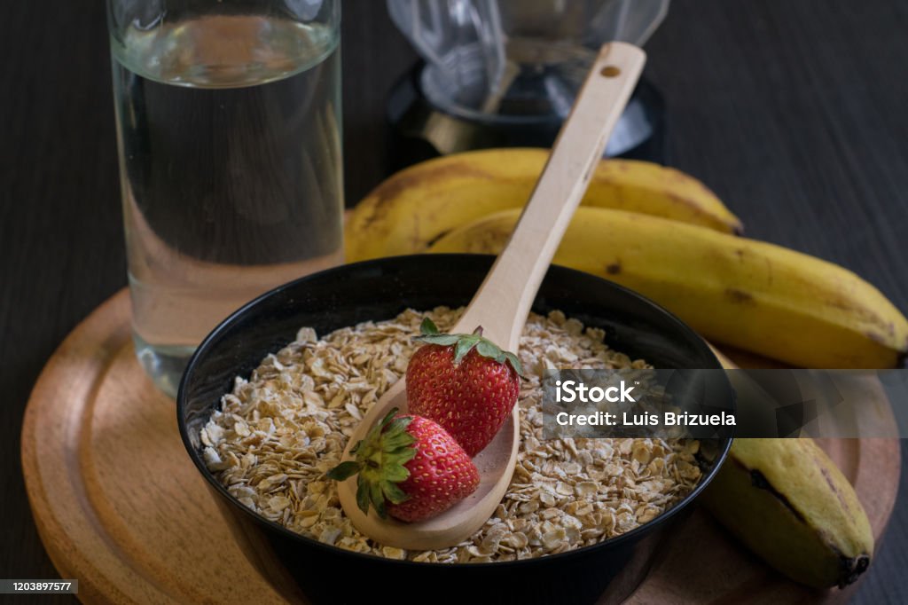 Strawberries oatmeal and bananas with a bottle of water oatmeal bowl with a wooden spoon with two strawberries with a bottle of water and bananas around and a blender jar back, on top of a round wooden board Argentina Stock Photo