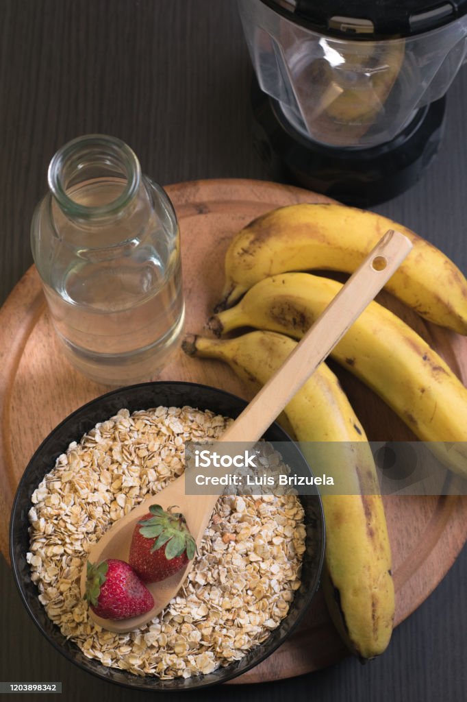 Strawberries oatmeal and bananas with a bottle of water top view of a wooden spoon with two strawberries above a bowl full of oatmeal with a bottle of water and bananas around with a blender jar back Argentina Stock Photo