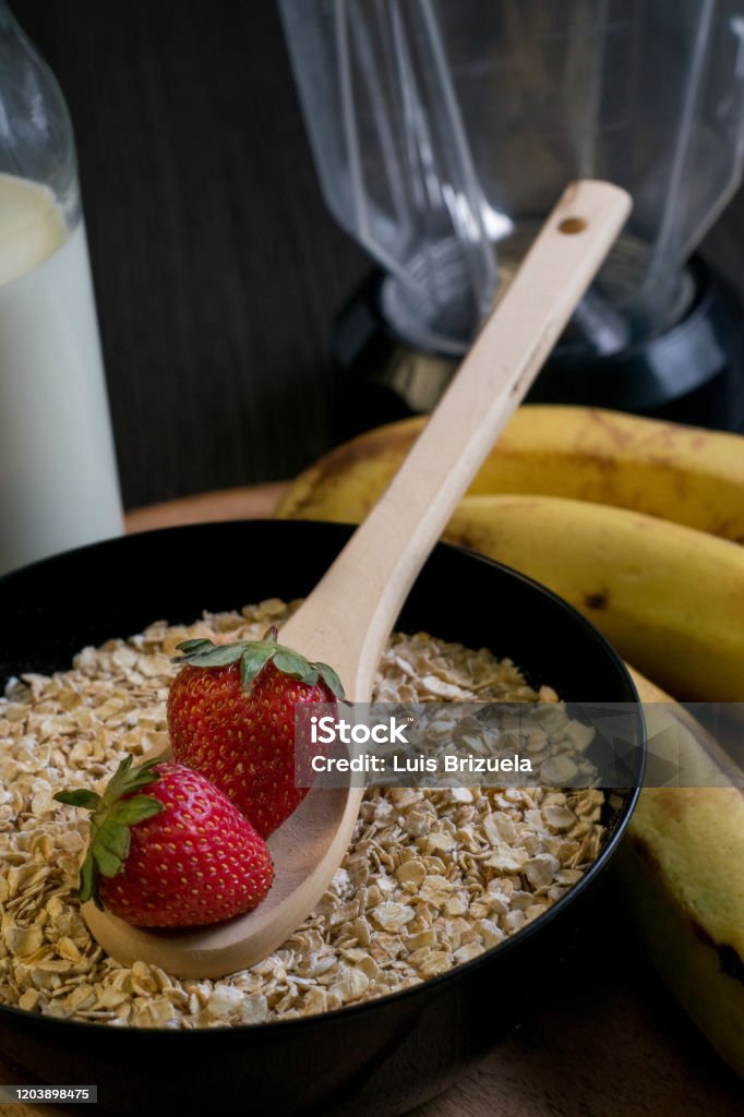 Strawberries oatmeal and bananas with a bottle of milk Close-up of two strawberries in a wooden spoon on a bowl full of oatmeal with bananas and a bottle of milk in the background Argentina Stock Photo
