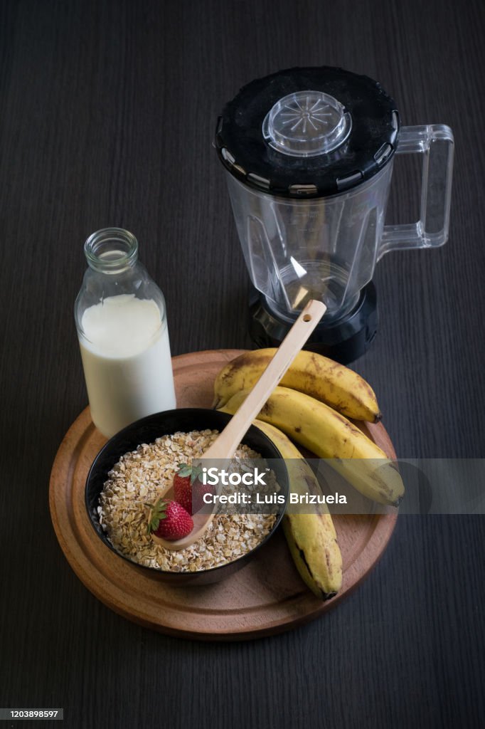 Strawberries oatmeal and bananas with a bottle of milk oatmeal bowl with a wooden spoon with two strawberries with a bottle of milk and bananas around and a blender jar back, on top of a round wooden board Argentina Stock Photo