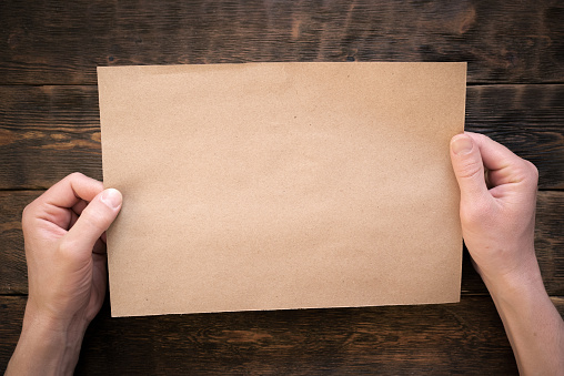 Blank brown paper page in male hands on wooden desk flat lay background.