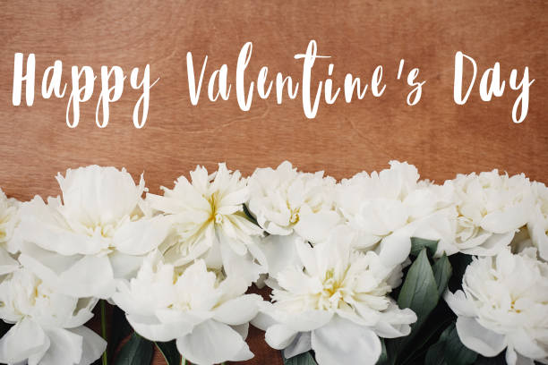 istock Happy Valentine's Day text on stylish white peony flat lay on rustic wooden table. Greeting card mockup. White peonies rural still life, top view. 1203903072
