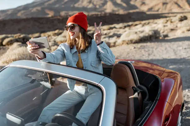 Young woman traveling by convertible car on the picturesquare road of the desert valley, photographing or vlogging on mobile phone