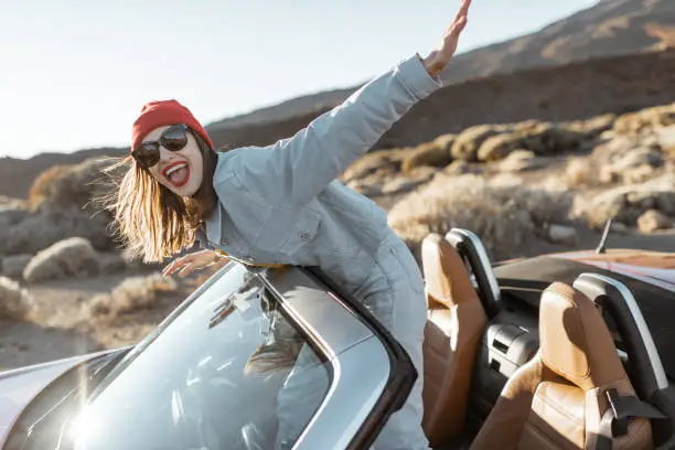 Lifestyle portrait of a playful woman having fun during a road trip, raising hands out of the convertible car while driving on the desert valley on a sunset