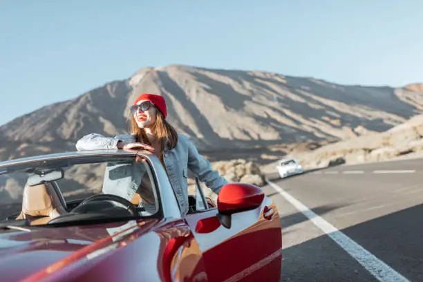 Young woman enjoying road trip on the desert valley, standing near the convertible car on the roadside with volcano on the background