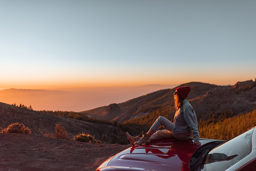 Landscape view on the roadside above the clouds with woman enjoying beautiful sunset, sitting on the convertible sports car