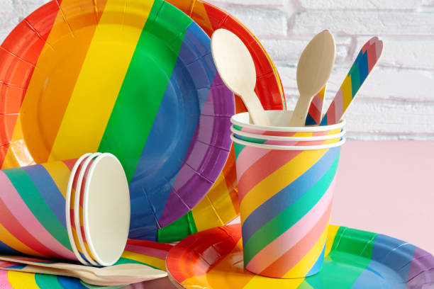 Rainbow colored disposable eco tableware on pink color background over white brick wall. Birthday party accessories,picnic utensil concept.