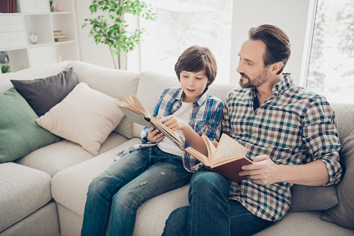 Portrait of two nice attractive focused cheerful smart clever guys dad and pre-teen son sitting on couch reading interesting novel story, book in light white modern style interior living-room