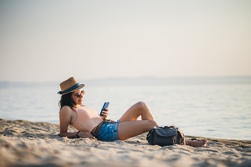 Young young caucasian cute woman sitting at sandy beach at sunset and texting on phone.