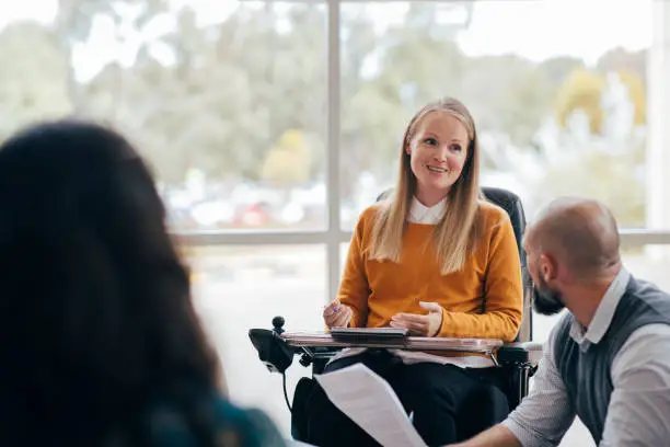 A woman looks happy and confident as she leads a group discussion at her place of work. She is a wheelchair user and has Muscular Dystrophy.