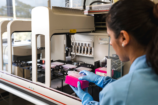 An out of focus Latin female scientist stands over a large sampling machine in a laboratory.