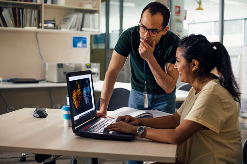 A male and female scientist each look at some results from a diagram displayed on a laptop. He is focussed and she is smiling as they work together to discuss their findings.