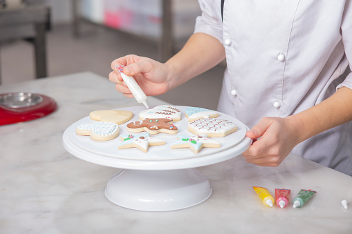 Female sugar baker is decoration christmas cookies with pink icing or frosting, workspace