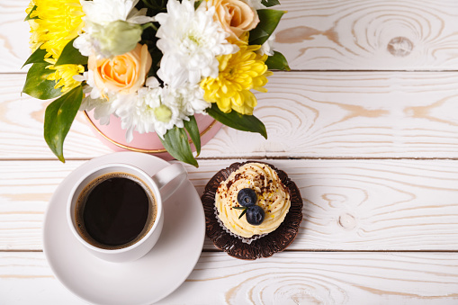 Cup of coffee, a cupcake with whipped cream and blueberries, a bouquet of spring flowers on a white background of a wooden table, top view. Romantic love background, mother's day greeting card