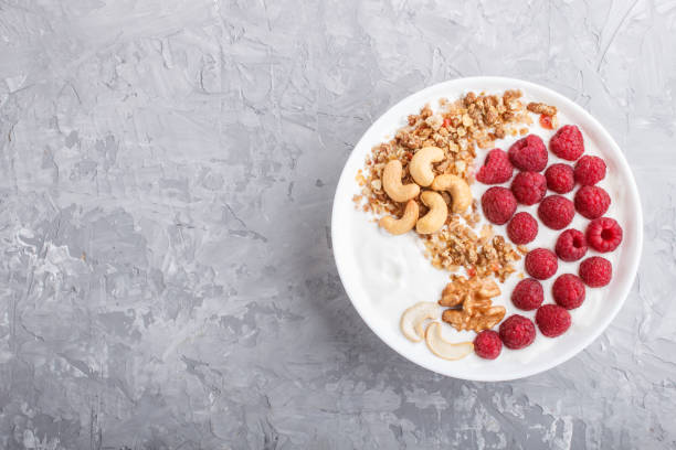 istock Yoghurt with raspberry, granola, cashew and walnut in white plate on gray concrete background and linen textile. top view, flat lay, copy space. 1203957827