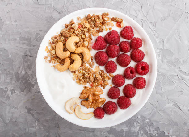 istock Yoghurt with raspberry, granola, cashew and walnut in white plate on gray concrete background and linen textile. top view, flat lay, close up. 1203960153