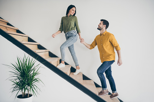 Let's explore this place. Full size profile photo of handsome guy and his pretty, lady walking to bedroom go up stairs in just bought modern flat indoors wear casual clothes