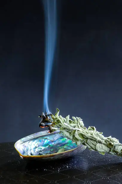 Photo of White sage smudge stick on dark background. Burning / smudging sage on abalone shell. Native American tradition for cleansing negative energy.