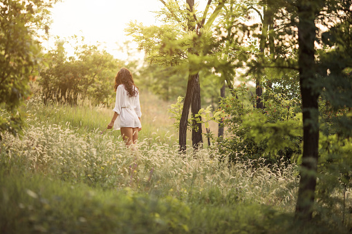 Beautiful woman on nature. Young lady on meadow in forest during sunset, wear summer white dress. Back view of attractive young girl outdoor.