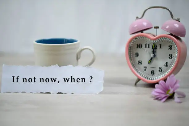 Inspirational quote - If not now, when. With a cup of coffee, pink clock alarm table and purple daisy flower on white wooden table background. Motivation words on paper note with motivating text question.