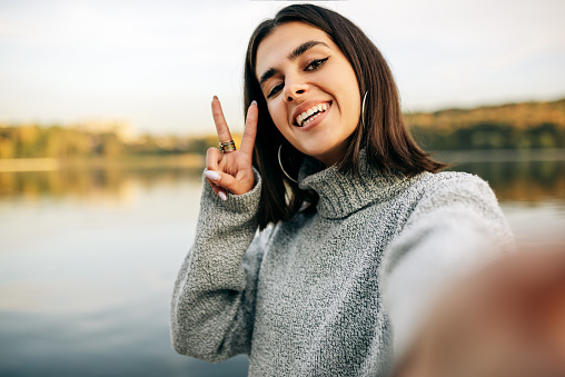 Pretty girl smiling broadly, wearing sweater making self portrait and showing peace gesture. Beautiful happy young female taking selfie on her device outdoor against nature background