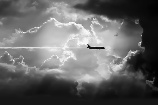 Black and white image of silhouette aircraft flying direct form bright sky through storm clouds in summer tropical season.