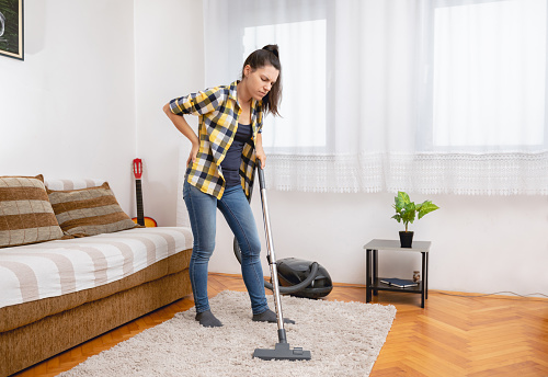 Young woman with backache vacuuming the apartment.